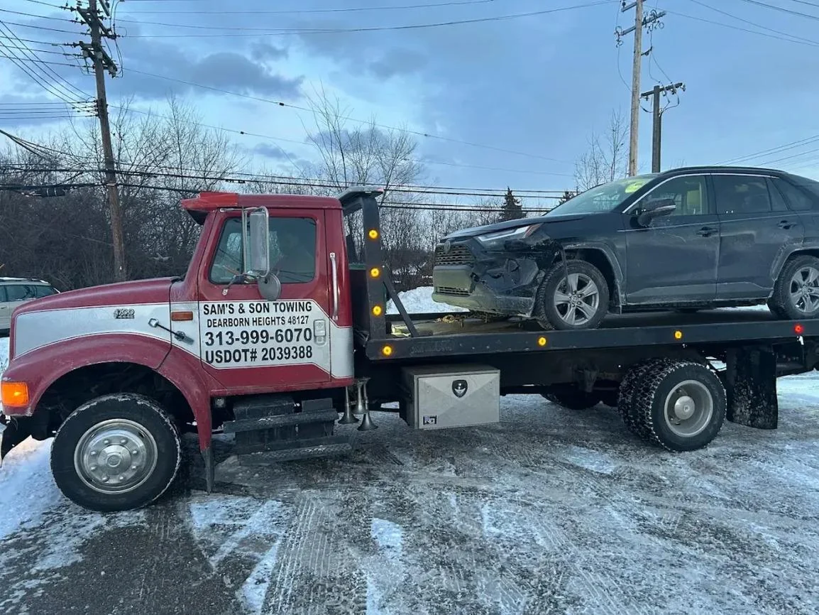 Sam's & Son flatbed carrying a dark SUV on a snowy day